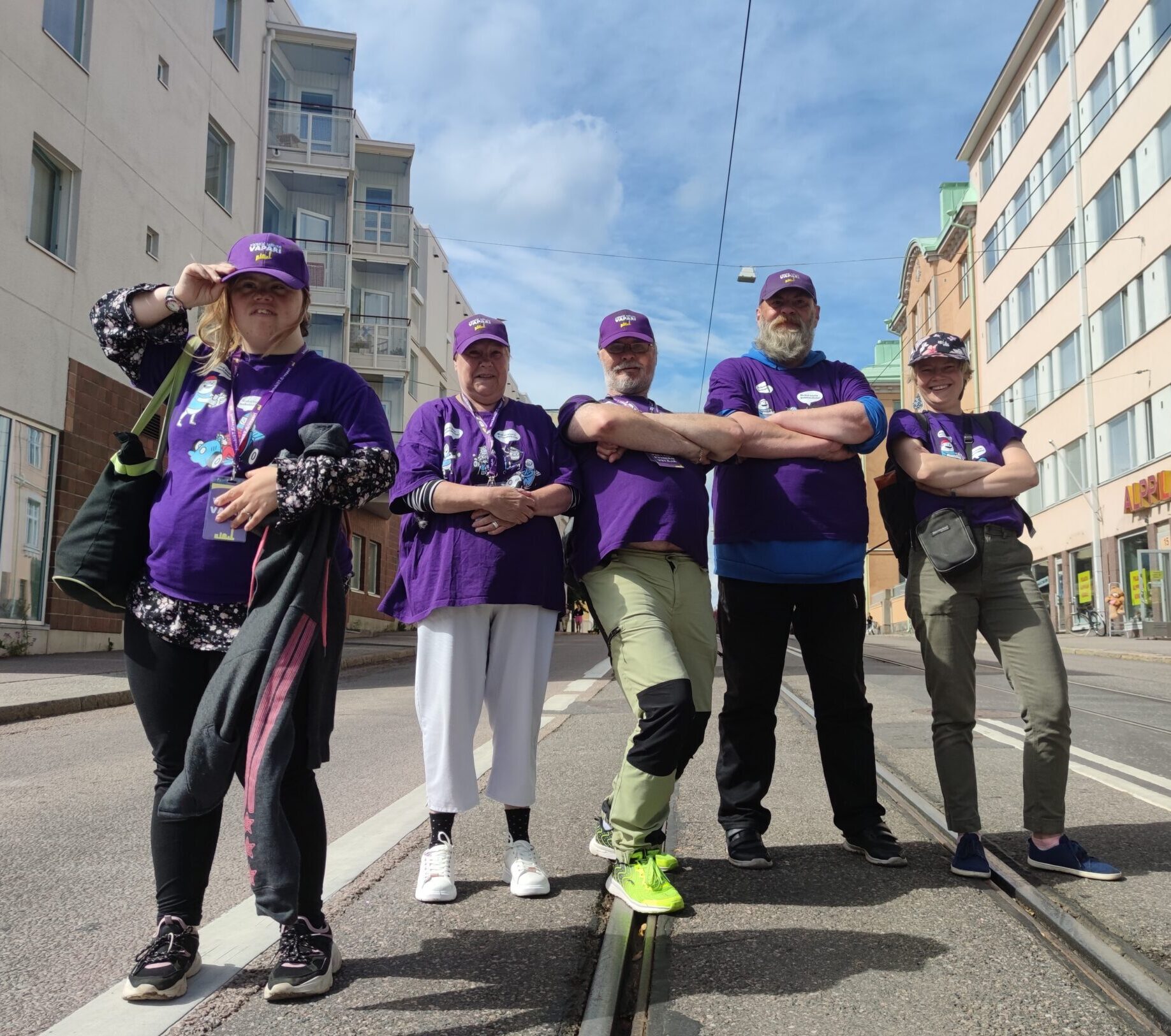 Viisi ihmistä seisoo kaupungin kadulla, katsoo kameraa ja hymyilee. Kaikilla on violetit paidat ja lippikset. Takana on poutapilvinen taivas.