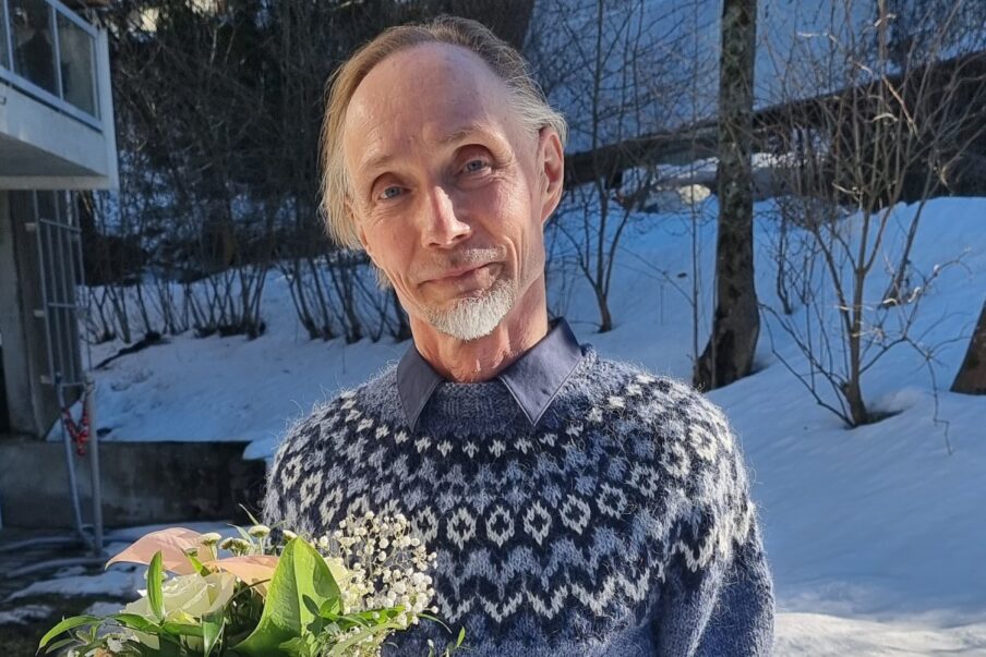 Mies, kukkakimppu kädessä, kirjoneulevillapaita päällä. takana hämärä talvimaisema.