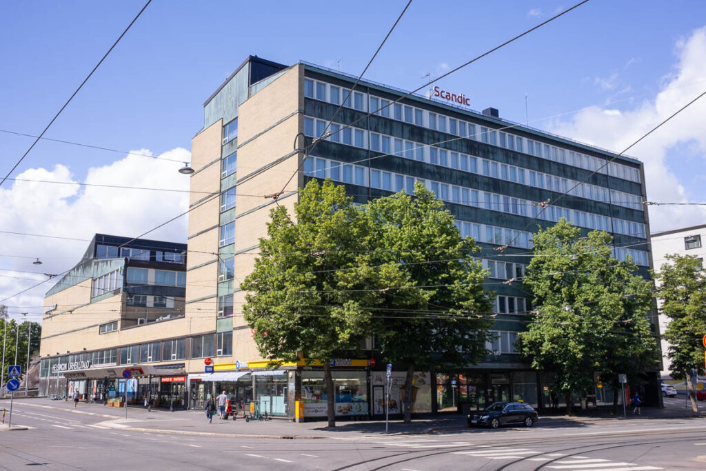 Kahdeksankerroksinen rakennus, jonka kaksi sivua näkyvät. Oikeanpuolimmainen sivu on väritykseltään harmaa ja joka kerroksessa on seinän mitan verran ikkunoita. Toinen sivu on keltaisesta tiilestä ja siinä on vähän ikkunoita. Talon edustalla on lehtipuita, jalkakäytävä ja raitiovaunukiskot.