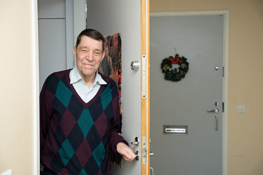 Mies seisoo asunnon oviaukolla, katsoo kohti ja hymyilee.