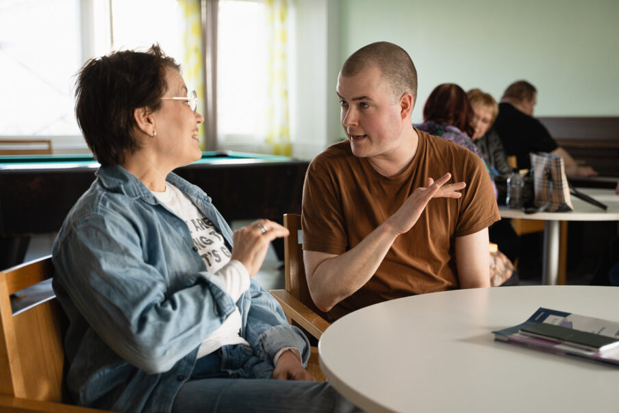 Mies ja nainen istuvat pöydän ääressä ja keskustelevat käsillä elehtien. Taustalla on lisää pöytiä, joissa istuu ihmisiä.