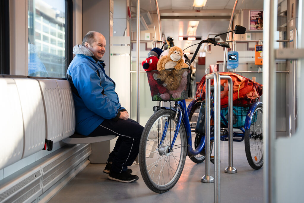 Mies istuu junassa ja katsoo hymyillen kolmipyöräistä sähköpyörää. Pyörän etukorissa on pehmoeläimiä.