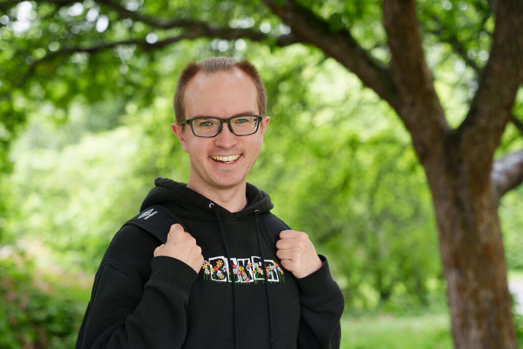 Mies, jolla on silmälasit katsoo kohti kameraa ja hymyilee. Hänen ympärillään on vihreää metsää.