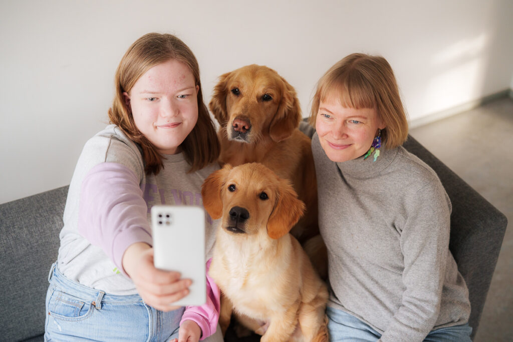 Kaksi ihmistä ja kaksi koiraa istuvat sohvalla ja ottavat selfietä.