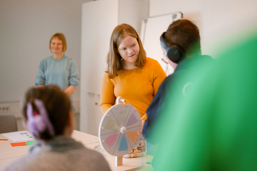 Ryhmä ihmisiä osallistuu pöydän ääressä toimintaan, jossa käytetään värillistä onnenpyörää. Pöydällä on papereita ja kyniä, ja taustalla näkyy valkotaulu ja kaappi.