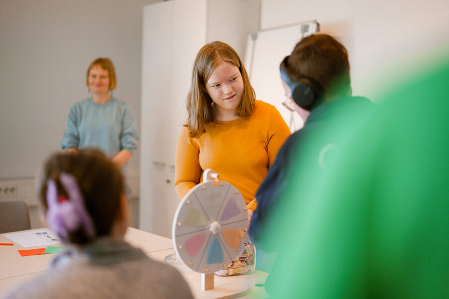 Ryhmä ihmisiä osallistuu pöydän ääressä toimintaan, jossa käytetään värillistä onnenpyörää. Pöydällä on papereita ja kyniä, ja taustalla näkyy valkotaulu ja kaappi.