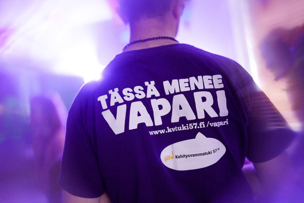 Henkilön selkäpuolelta otettu kuva. Paidassa teksti: tässä menee vapari www.kvtuki57.fi/vapari ja Kehitysvammatuki 57 ry:n logo.