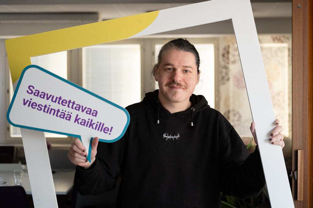 Kuvassa harjoittelija Anton hymyilee ja pitää kädessään Saavutettavaa viestintää kaikille kylttiä