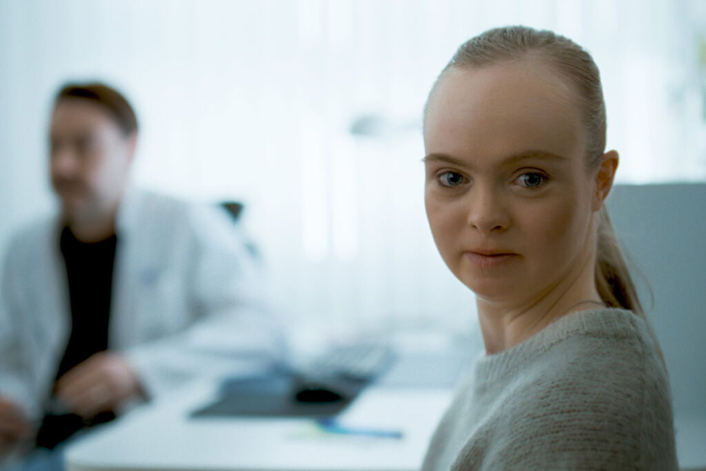 Nuori nainen on lääkärissä ja katsoo kohti. Taustalla lääkäri puhuu naisesta poispäin.