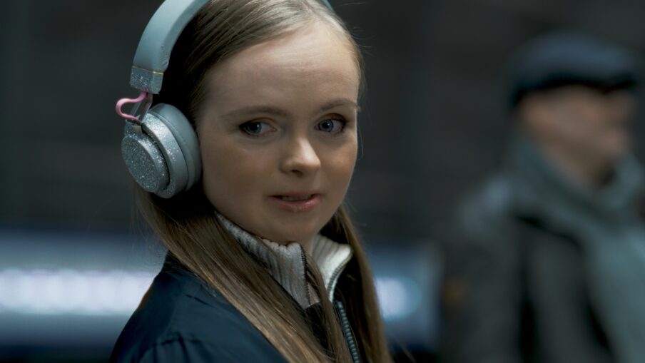 Nainen katsoo kohti. Hänellä on päässään kuulokkeet. Taustalla näkyy henkilö, joka katsoo toiseen suuntaan.