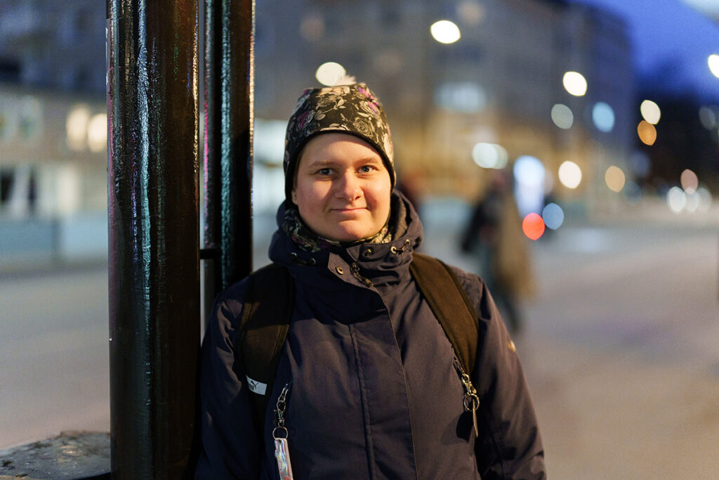 Nainen on ulkona, nojaa tolppaan ja katsoo kohti kameraa.