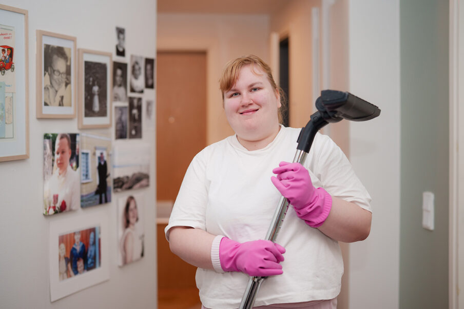 Nuori nainen hymyilee kameralle. Hänellä on käsessä vaalanpunaiset siivoushanskat ja imuri.