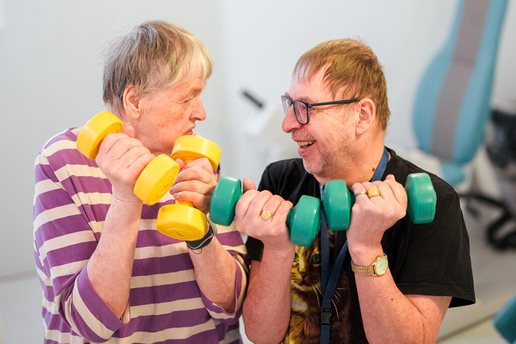 Kaksi ikääntynyttä ihmistä nostelee käsipainoja ja hymyilevät toisilleen.