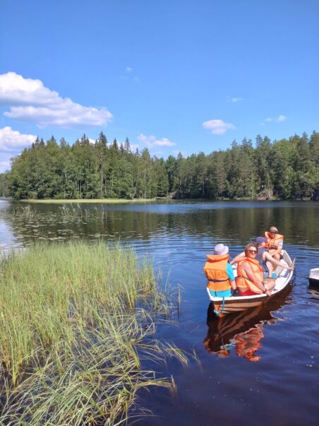 kesäinen järvimaisema ja vene. Veneessä neljä henkilöä pelastusliiveissä.