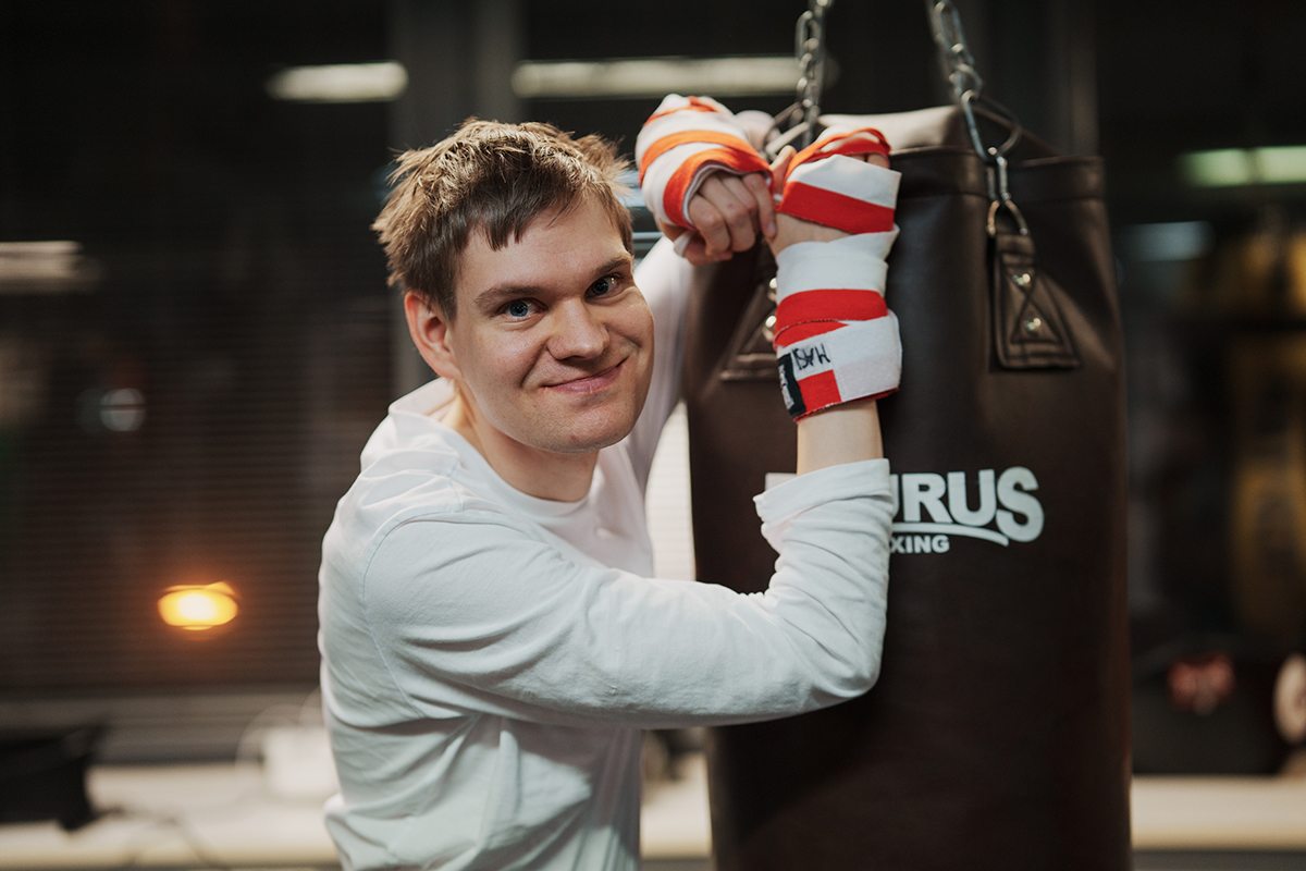 Hymyilevä ihminen, jolla on nyrkkeilyhanskat kädessä nojaa nyrkkeilysäkkiin.