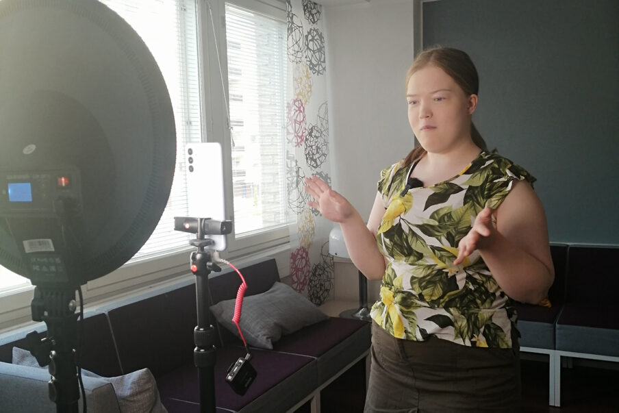 Nuori nainen puhuu telineeseen asetettuun puhelimeen. Vieressä on kuvausvalo, joka valaisee puhujan kasvoja.