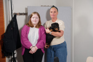 Kaksi henkilöä katsoo kameraan. Toisella on mäyräkoira sylissään.