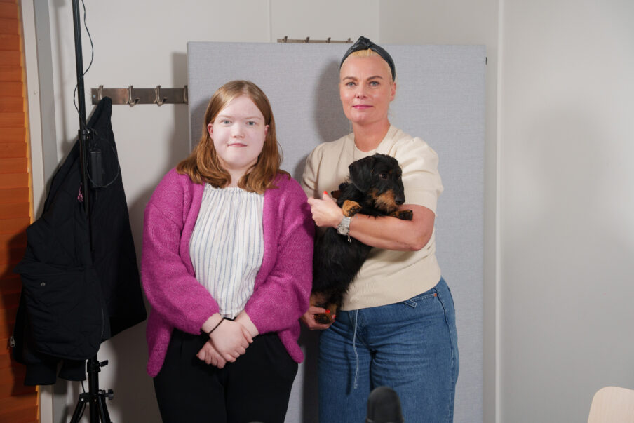 Kaksi henkilöä katsoo kameraan. Toisella on mäyräkoira sylissään.